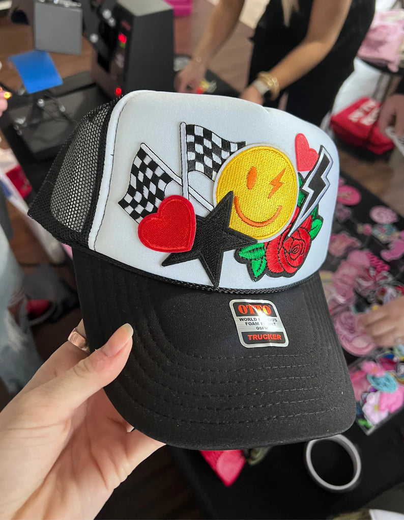 A person is holding a black and white baseball cap with various colorful and decorative patches, including a smiley face, hearts, and flags.